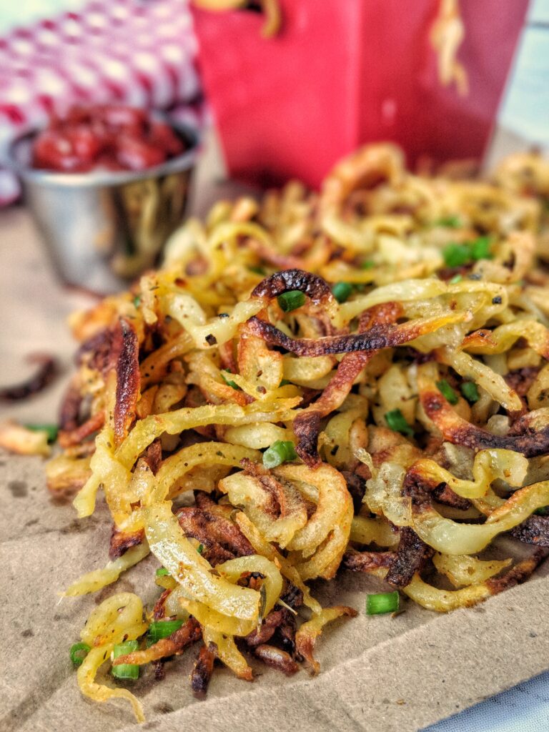 Crisped curly fries garnished with green onions are piled high on a brown piece of paper alongside a metal ramekin of ketchup. Seen blurred in the background is a red cardboard box of curly fries also placed on the piece of paper.
