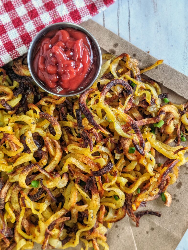 Thin crispy curly fries are scattered across a piece of brown paper alongside a ramekin of ketchup.