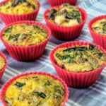Arranged on a light blue plaid cloth are oven and air fryer safe red silicone muffin filled with bright yellow cooked egg loaded with dairy-free nutritional yeast, sausage, potatoes, onion and kale.