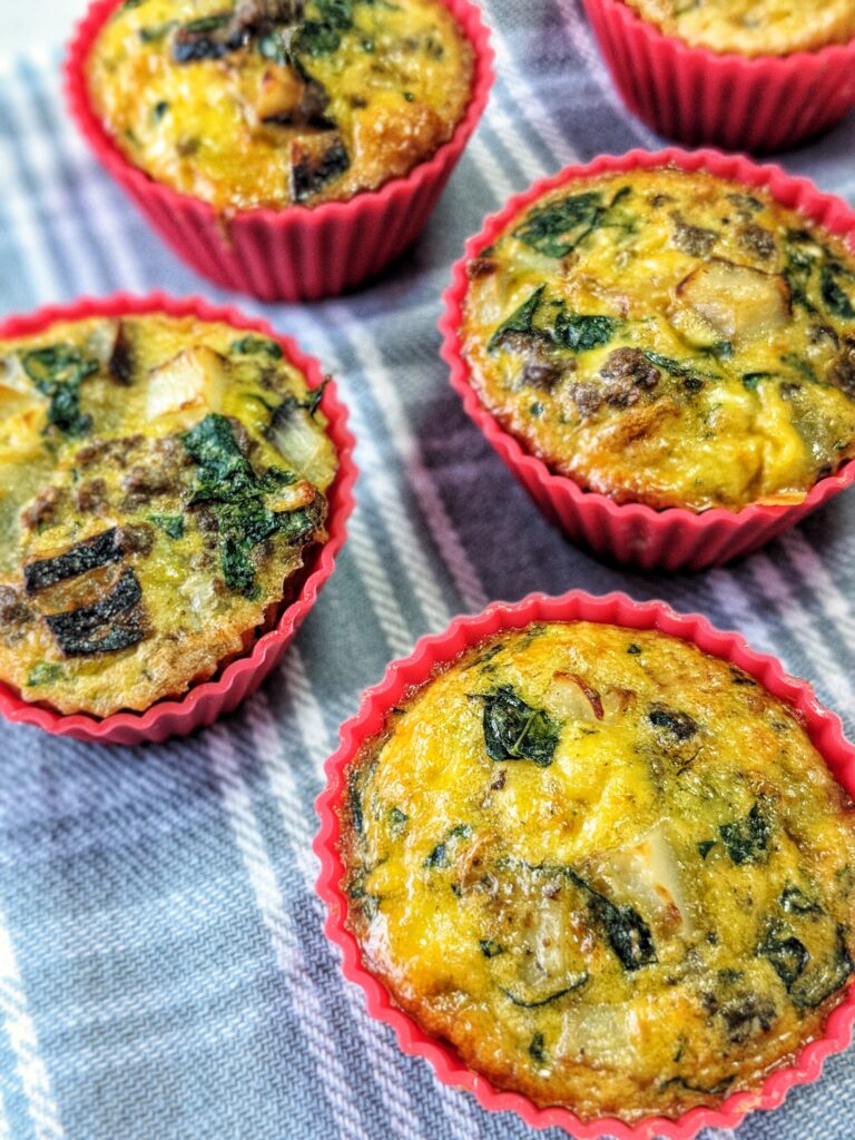 5 red silicone baking cups are scattered across a pale blue tablecloth. Each cup is filled with egg breakfast muffins loaded with sausage, potatoes, onions, kale and nutritional yeast. 