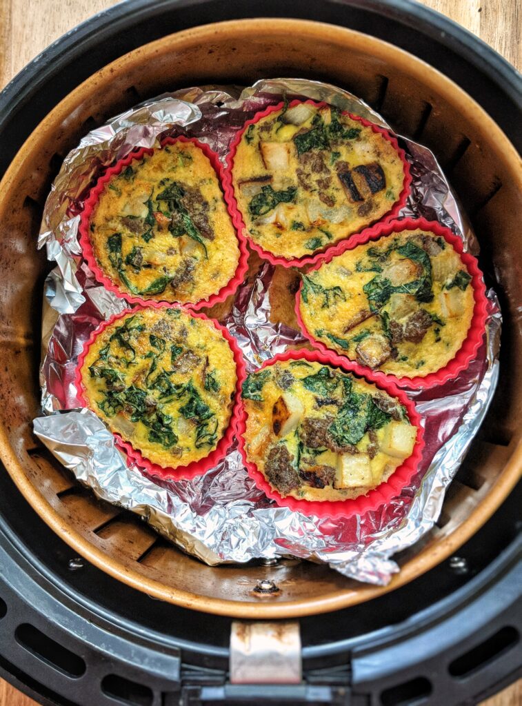 5 red silicone baking cups are arranged in an air fryer basket. In each cup is a cooked breakfast cup loaded with eggs, sausage, potatoes, onions, kale and nutritional yeast.