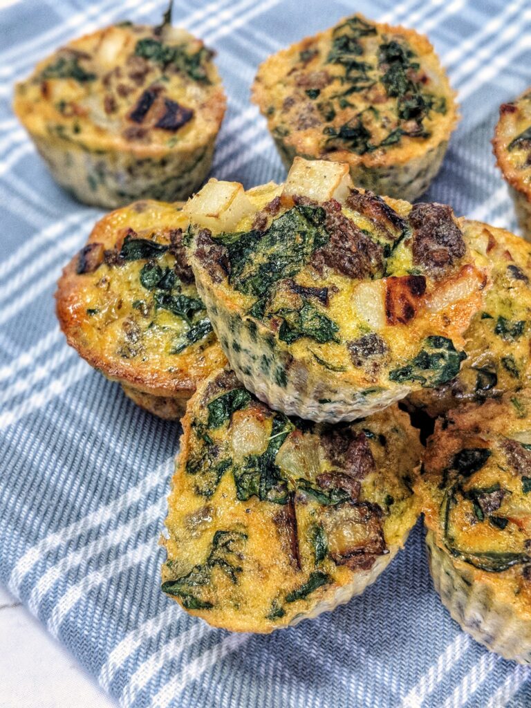 Scattered on a light blue plaid cloth are bright yellow cooked egg muffins loaded with dairy-free nutritional yeast, sausage, potatoes, onion and kale.