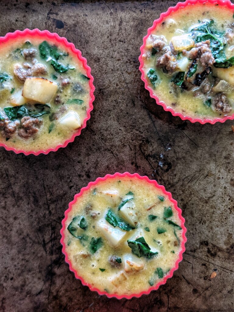 3 red silicone baking cups are arranged on a baking sheet. Each cup is filled with a mixture of raw egg, sausage, potatoes, onions, kale and nutritional yeast.