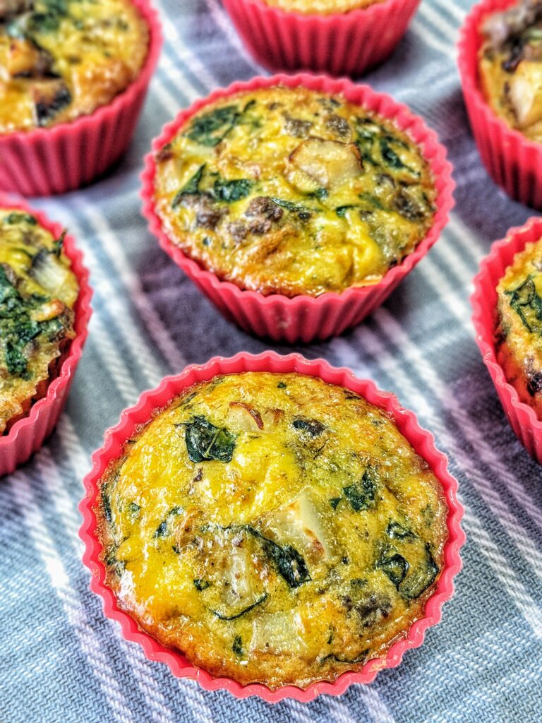 Arranged on a light blue plaid cloth are oven and air fryer safe red silicone muffin cups filled with bright yellow cooked egg loaded with dairy-free nutritional yeast, sausage, potatoes, onion and kale.