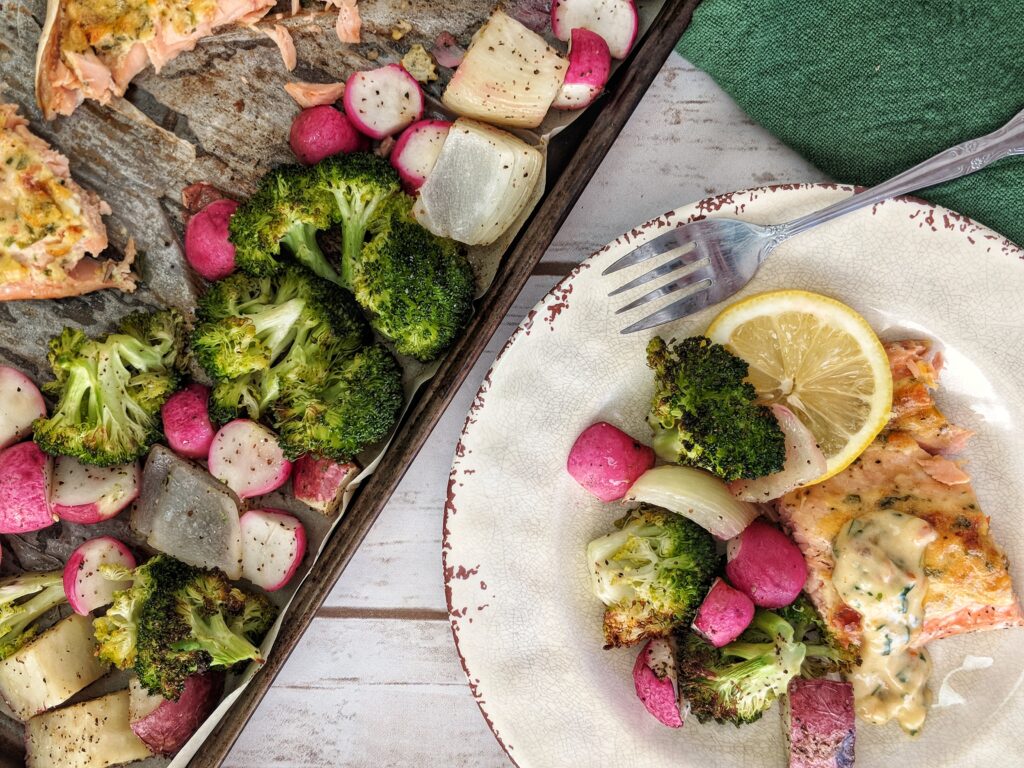 A portion of baked salmon fillet and roasted vegetables is plated with a lemon wheel and a dollop of sun-dried tomato aioli. The remaining salmon and veggies is still on the sheet pan.