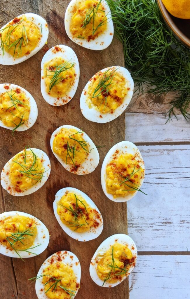 A dozen perfectly presented Creamy-Fennel Deviled Eggs sit on top of a wooden plank. Each egg is garnished with a few sprigs of fresh fennel fronds and a sprinkle of paprika.