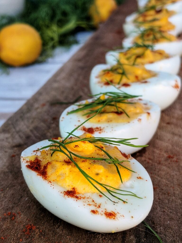Neatly arranged on a piece of rustic wood are several Paleo Creamy-Fennel Deviled Eggs. The bright yellow yolks are highlighted by a sprinkle of fresh fennel fronds and paprika.