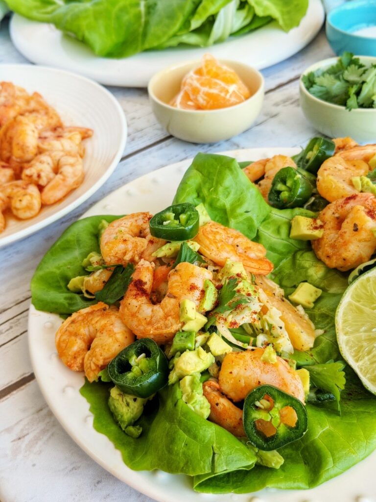 Displayed on a bright white plate are two Blackened Shrimp Lettuce Wraps topped with diced avocado, jalapeno slices and cilantro.
