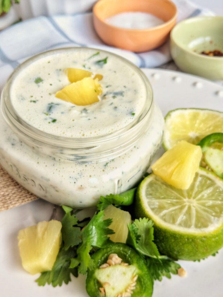 Scattered around a small mason jar of creamy jalapeno pineapple aioli are pineapple chunks, lime wheels and cilantro sprigs. Pinch cups of sea salt and red pepper flakes are displayed in the background.