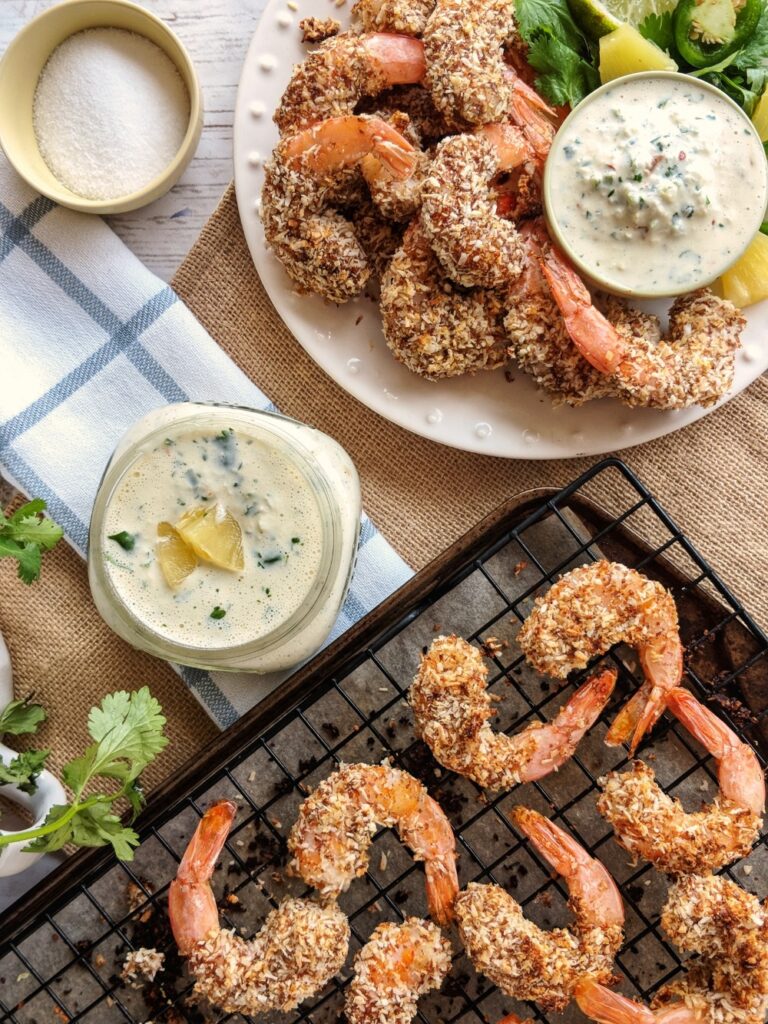 Golden baked Kombucha-Battered Coconut Shrimp are presented on a white plate with Pineapple Jalapeno Aioli and a garnish of lime, cilantro, jalapeno and pineapple. A baking sheet of finished shrimp is also presented alongside a mason jar of more Pineapple Jalapeno Aioli.