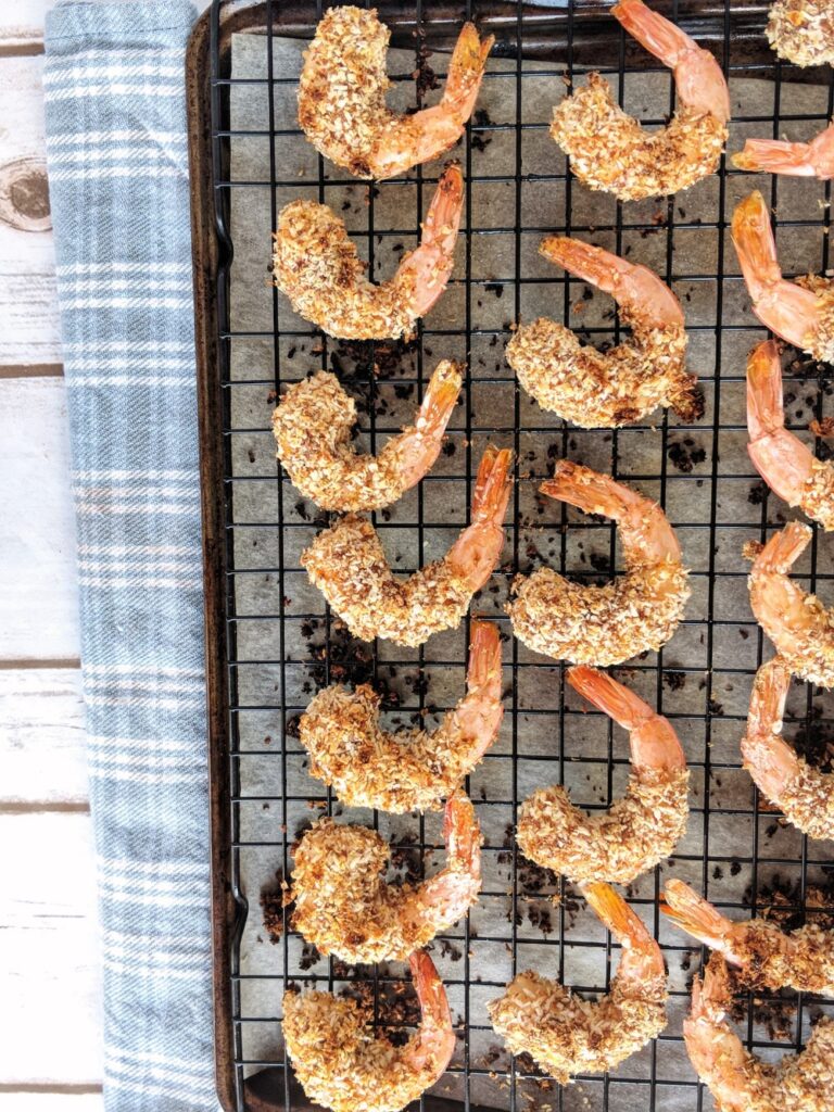 Perfectly toasted Kombucha-Battered Coconut shrimp are displayed fresh from the oven on a wire rack positioned above a parchment paper-lined baking sheet. The baking sheet is placed on top of a light blue plaid tea towel.