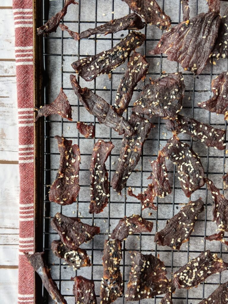 Tender, cooked pieces of Homemade Everything Bagel Beef Jerky are scattered on a cooling rack above a parchment paper-lined baking sheet.