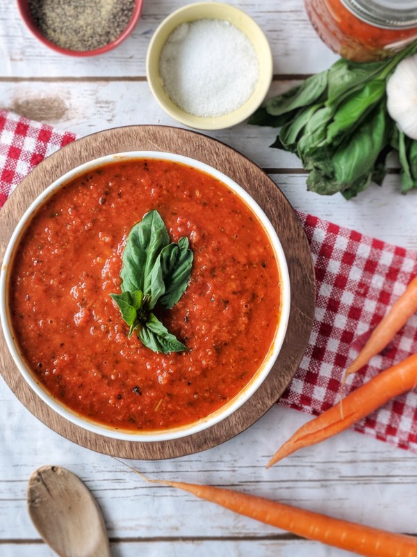 Vibrant red marinara sauce is served in a bowl above a wooden plate. Salt, pepper, fresh basil, garlic, carrots and a wooden spoon are scattered around the bowl of sauce.