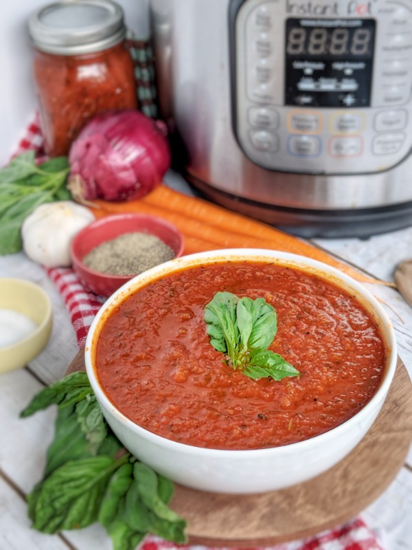 A vibrant bowl of fresh tomato sauce is presented in a white bowl above a wood plate. Fresh vegetables, herbs, a mason jar and an Instant Pot pressure cooker are seen in the distance.