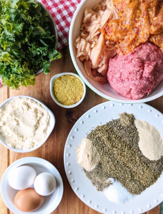 Overhead shot of the ingredients for Baked Italian Meatballs (kale, ground beef, chicken and pork sausage, nutritional yeast, almond flour, dried herbs and spices and eggs).