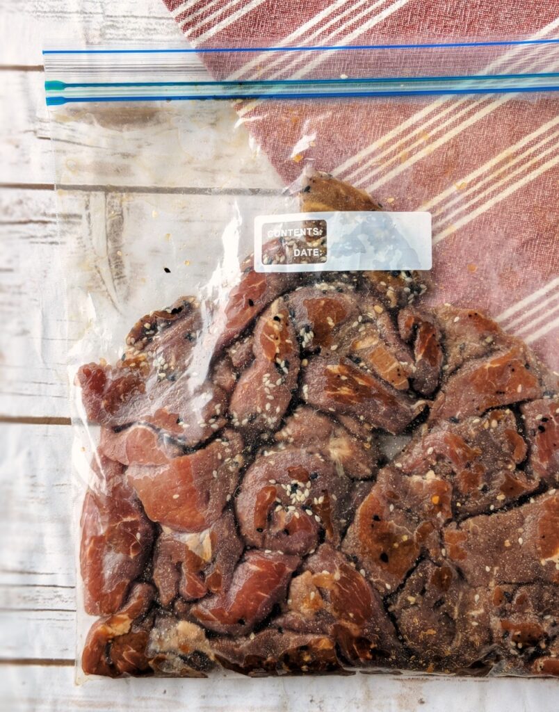 Thin strips of beef are marinating in coconut aminos, "everything bagel" seasoning and liquid smoke are placed in a plastic resealable bag.