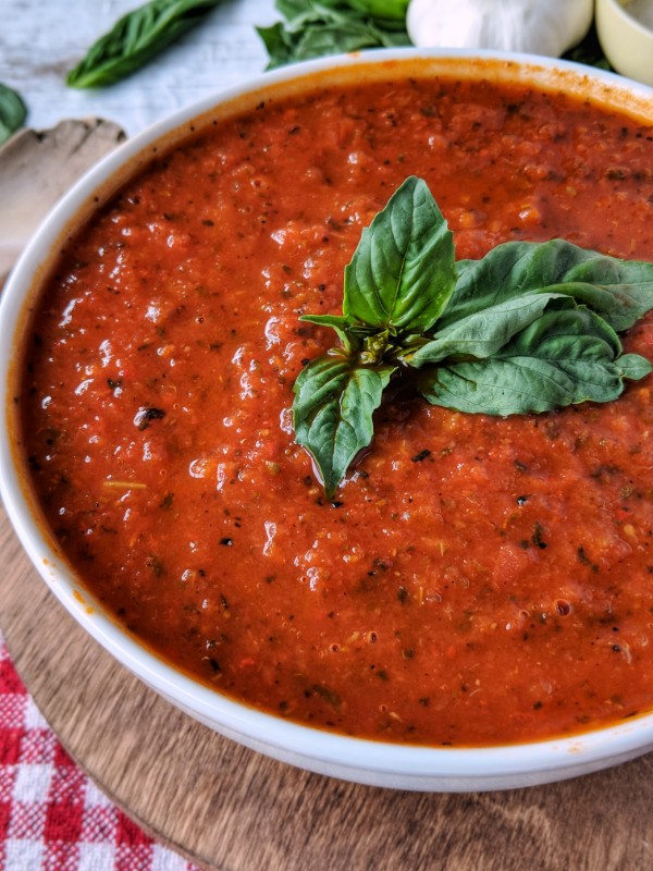 Perfect Instant Pot Paleo Marinara Sauce is presented in a bright white bowl placed on a red checkered tea towel. Fresh basil leaves, garlic, red onion, carrots and a pinch cup each of salt and pepper are placed around the bowl.
