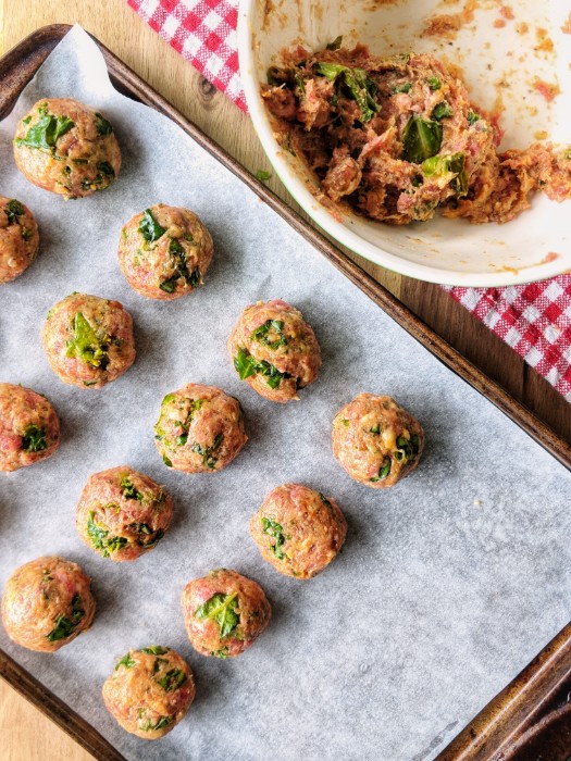 Uncooked Italian Meatballs are neatly placed in an even-layer on a parchment paper-lined baking sheet next to a large bowl with unrolled meatball mixture.