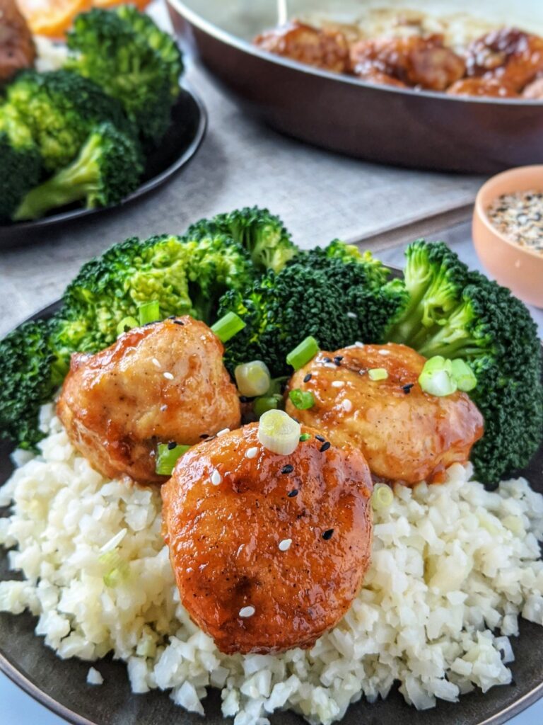 Plated on a matte black dish is a pile of fresh steamed broccoli, a bed of cauliflower rice and a serving of moist Paleo Meatballs glazed in a healthy Chinese Orange Chicken style sauce.