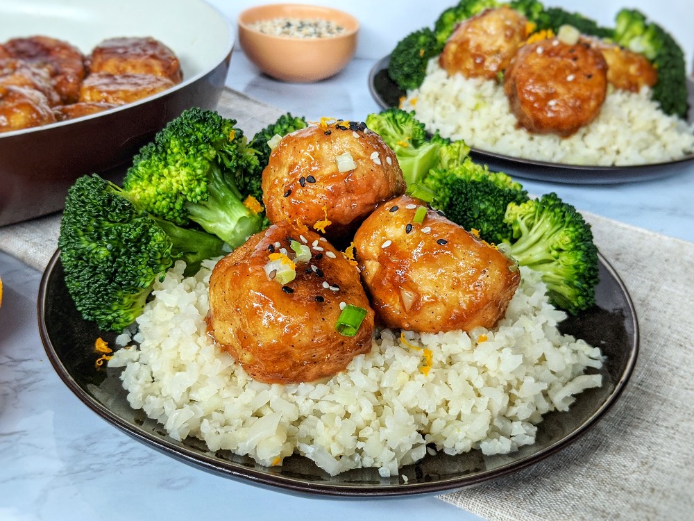 Three Gluten-free Orange Chicken Meatballs are plated on a black plate on a bed of fluffy cauliflower rice served alongside fresh steamed broccoli. The meal is garnished with sesame seeds, sliced green onions and fresh orange zest. The same plate and a skillet of cooked Whole30 Orange Chicken Meatballs is displayed in the background.