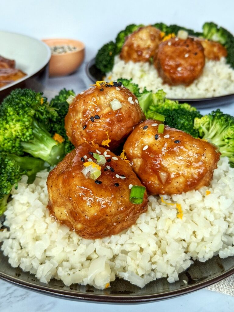 Three Orange Chicken Meatballs are plated on a black plate on a bed of fluffy cauliflower rice served alongside fresh steamed broccoli. The meal is garnished with sesame seeds, sliced green onions and fresh orange zest. The same plate and a skillet of cooked meatballs is displayed in the background.