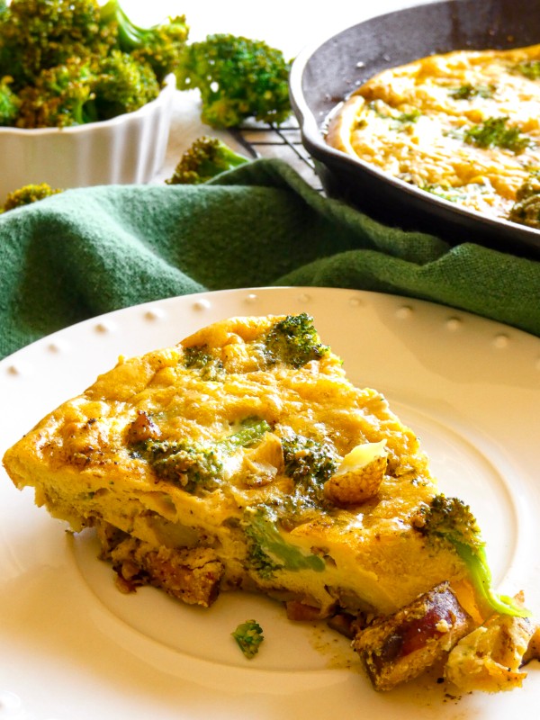 A slice of Smoked Sausage and Potato Frittata with Broccoli is served on bright white plate. The remaining frittata and a bowl of broccoli is blurred in the distance.