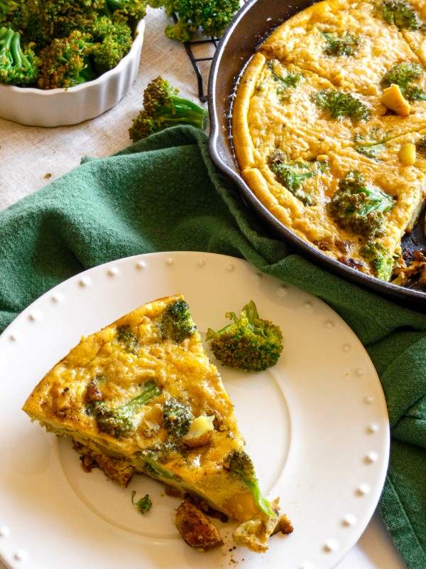 A slice of frittata loaded with meat and vegetables is presented on a small white plate alongside a cast-iron skillet of the remaining frittata and a bowl of broccoli.