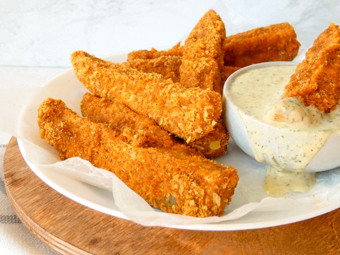 Crunchy breaded pickle spears are plated on top of one another. One of the spears is dunked in a small dish of creamy dairy-free ranch dressing.