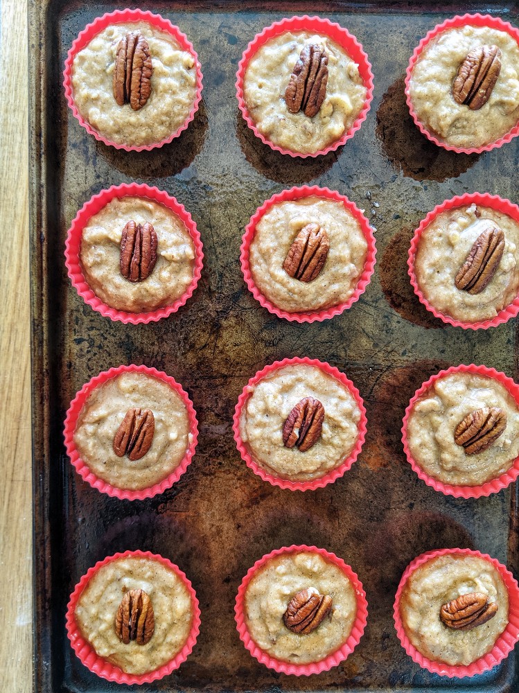 Placed above a baking sheet are 12 red silicone baking cups filled with Paleo Maple Pecan Banana Bread Muffin batter. A pecan half is pressed into the top of each cup of batter.